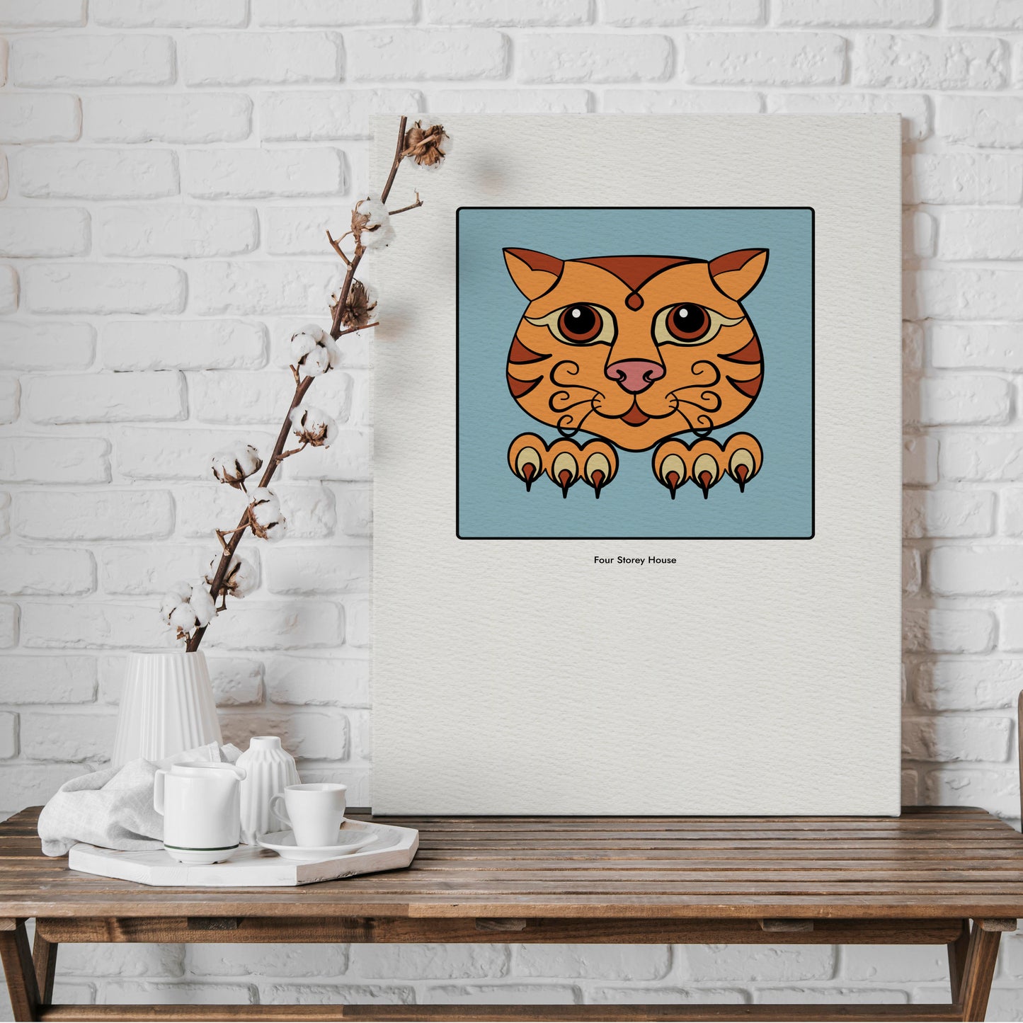 Original A2 Art Print - Ginger Cat featuring a stylized ginger cat face with large eyes and paws on a blue background, displayed on a textured cream canvas against a white brick wall.