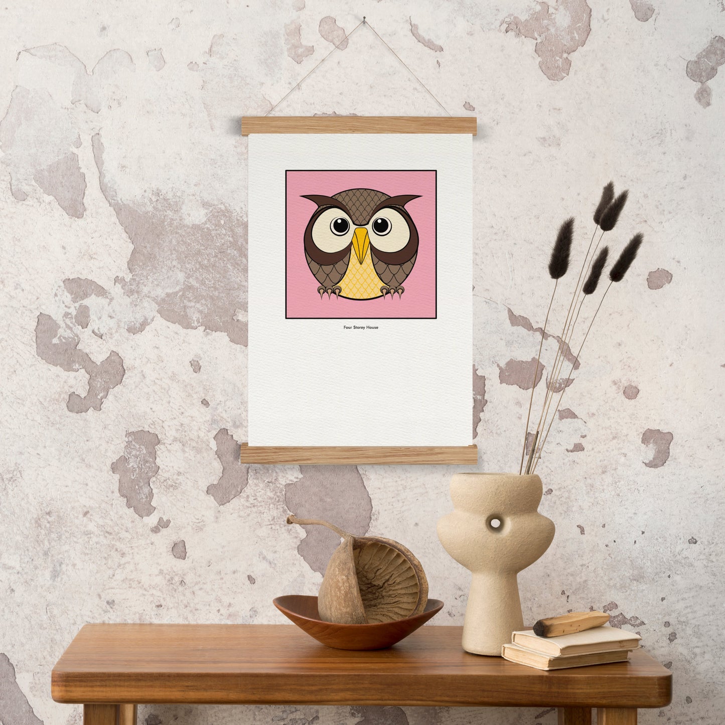 Original A2 Art Print - Owl featuring a stylized owl illustration in earthy tones on a pink background, displayed with wooden hanger on a textured wall above a wooden table with decorative items.
