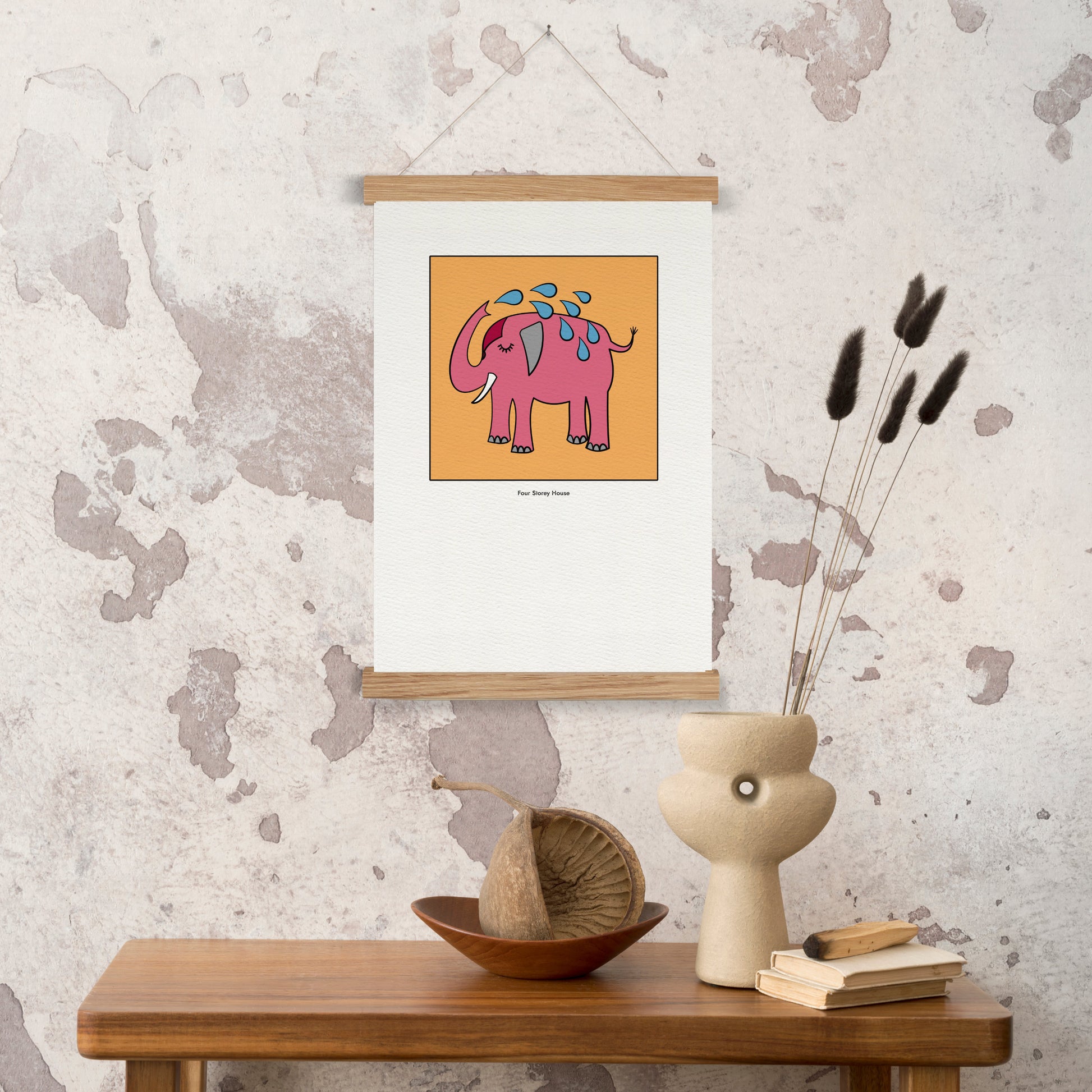Original A2 Art Print - Pink Elephant hanging on textured wall above wooden table with decorative vase, dried flowers, and books in a cozy, minimalist interior.